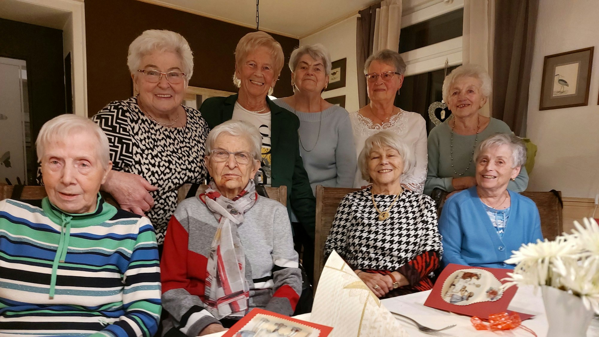 Vier Seniorinnen sitzen an einem Tisch, auf dem Weihnachtskarten liegen. Hinter ihnen stehen fünf weitere ältere Damen.