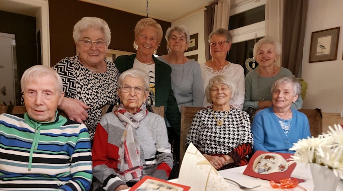 Vier Seniorinnen sitzen an einem Tisch, auf dem Weihnachtskarten liegen. Hinter ihnen stehen fünf weitere ältere Damen.&nbsp;