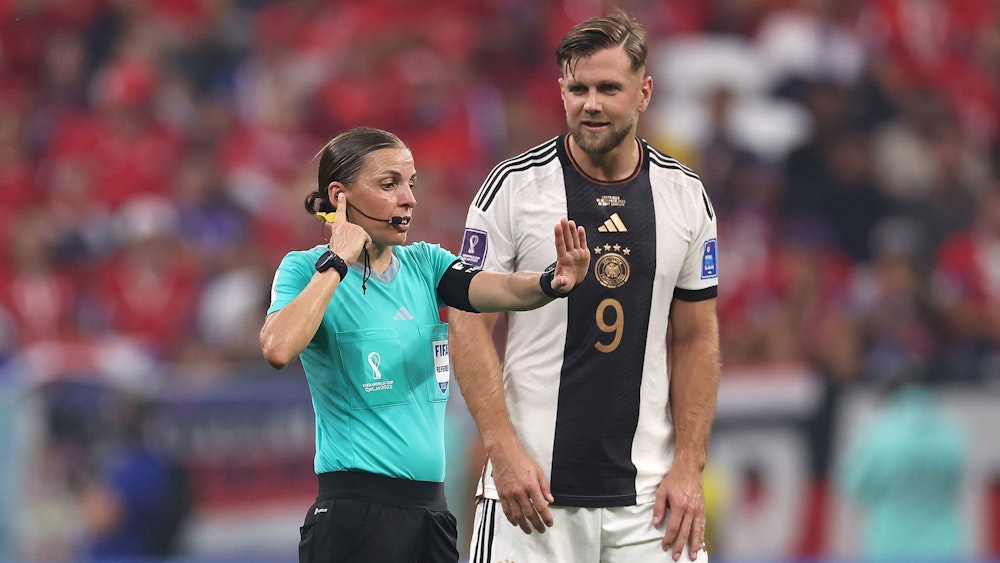 Schiedsrichterin Stéphanie Frappart greift sich bei der WM 2022 mit der Hand ans Ohr und kommuniziert mit dem VAR. Deutschland-Stürmer Niclas Füllkrug schaut interessiert.