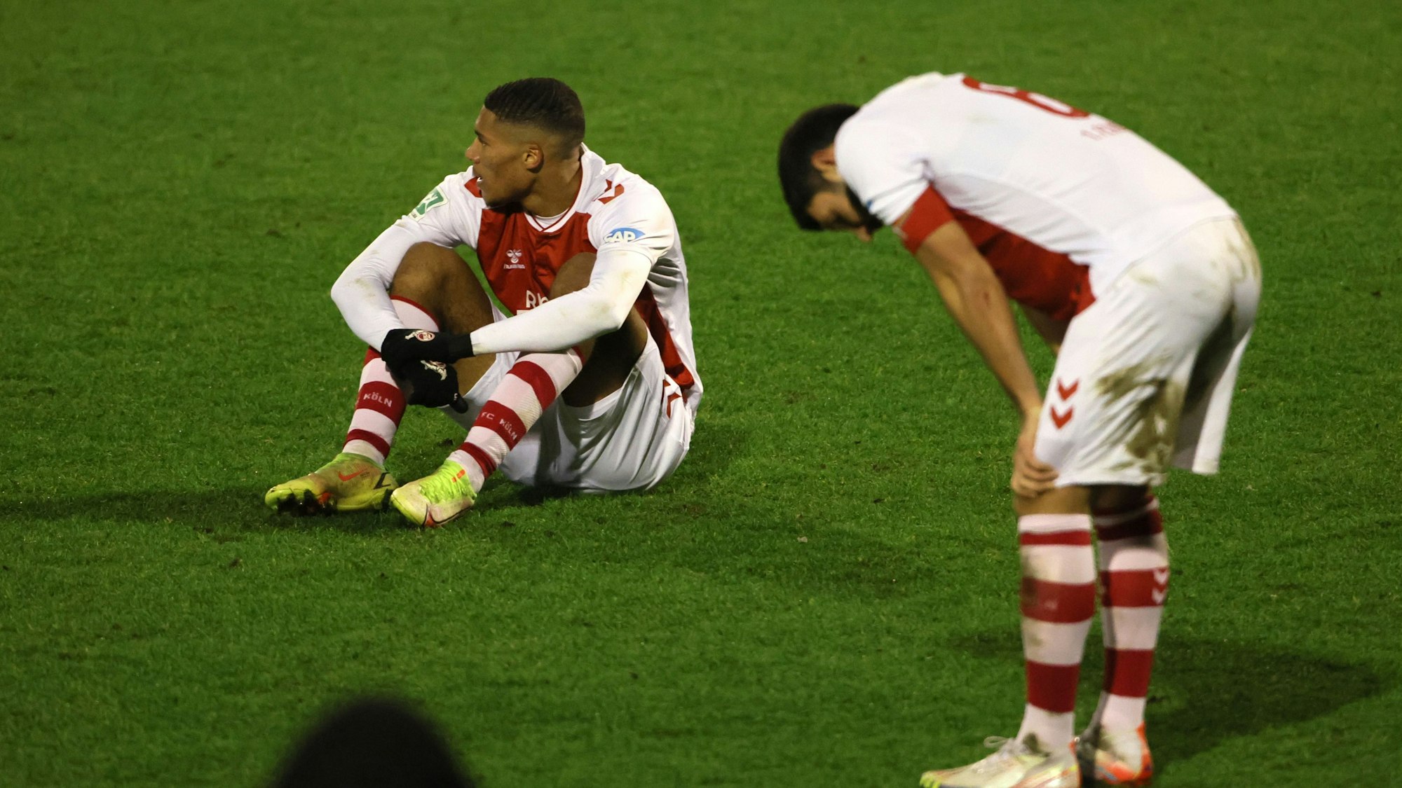 Ein Fußballspieler sitzt nach einer Niederlage enttäuscht auf dem Rasen, ein anderer stützt die Hände auf die Knie.