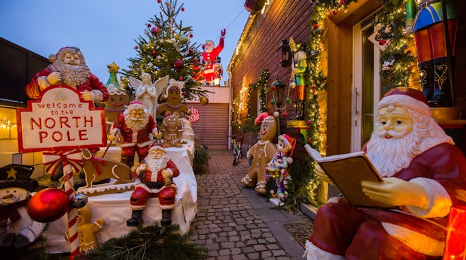 Mehrere Weihnachtsmänner, aber auch ein Engel und Lebkuchenfiguren stehen vor einem holzgetäfelten Haus. Auch ein geschmückter Weihnachtsbaum und ein Nikolaus auf einem Garagendach sind zu sehen.&nbsp;