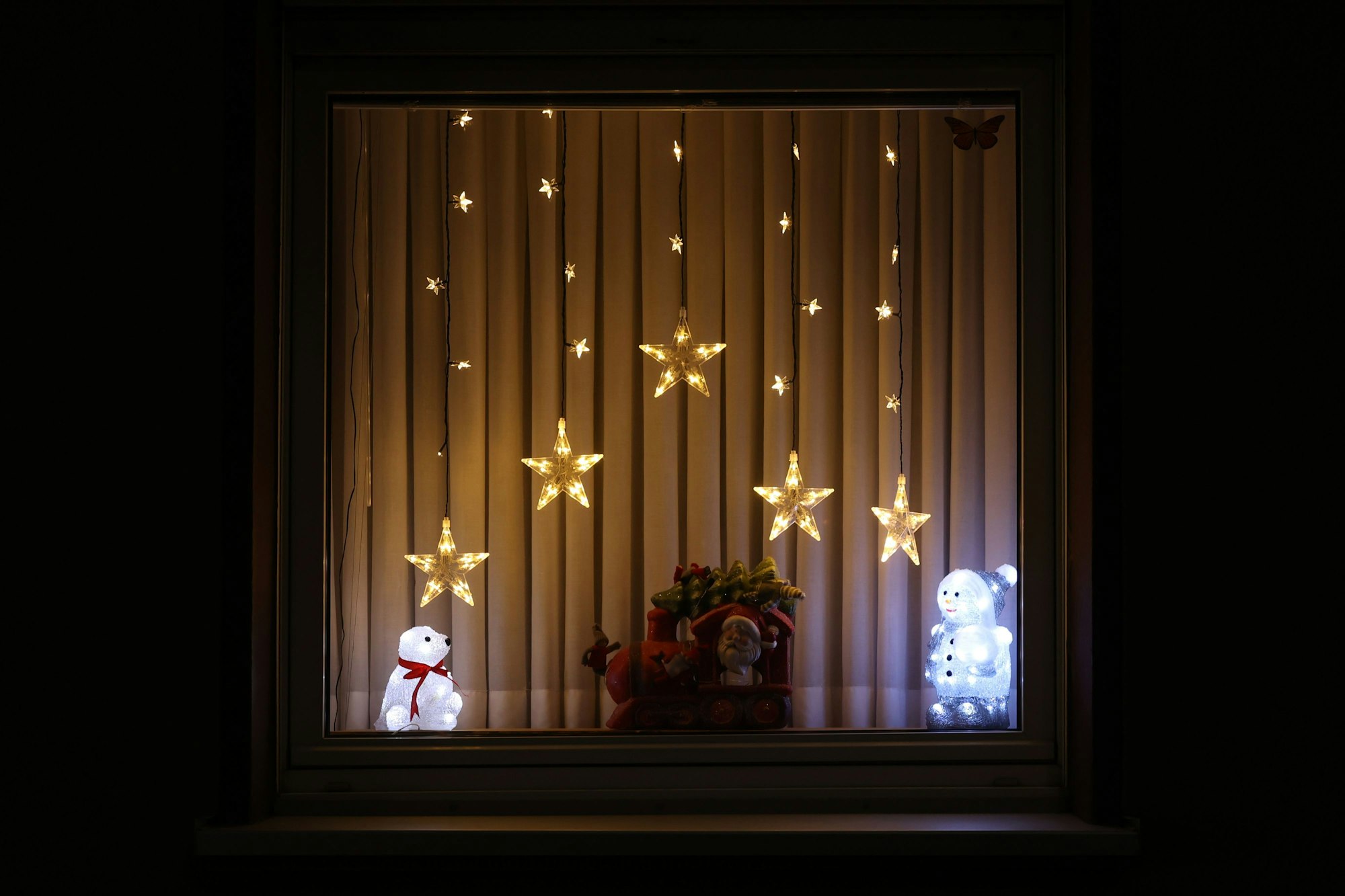 Leuchtende Sterne hängen in einem Fenster. Auf dem Fensterbrett steht ein leuchtender Eisbär sowie ein Schneemann.