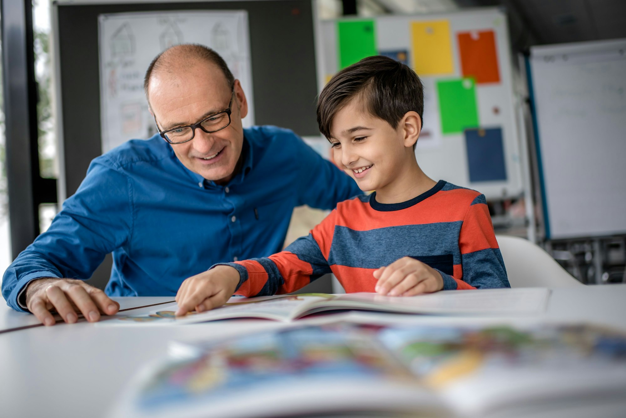 Mentor Andreas Endermann begleitet ein Kind beim Lesenlernen - das Bild zeigt die beiden, wie sie in einem Buch lesen, das vor ihnen auf dem Tisch liegt.