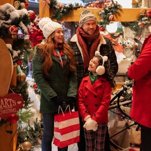 Eine Szene aus dem Film „Falling For Christmas“: Lindsay Lohan als Sierra, Chord Overstreet als Jake, Olivia Perez als Avy und Bus Riley als Kastanienverkäufer in weihnachtlicher Dekoration.