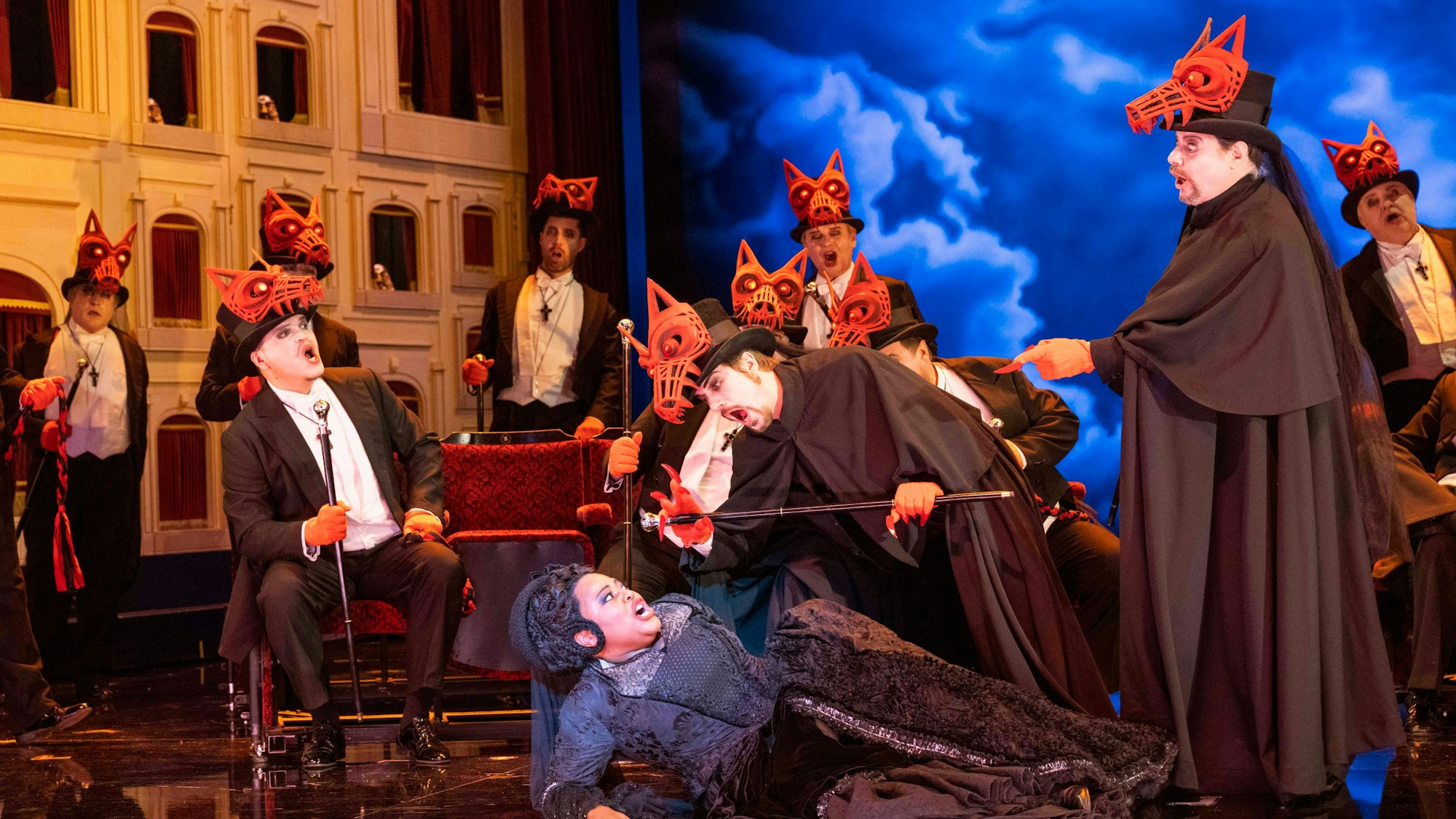 Eine Bühnenszene aus Verdis „Maskenball“: Eine Frau liegt am Boden, über ihr und um sie herum schwarz gekleidete Männer mit Wolfsmasken auf ihren Zylindern.