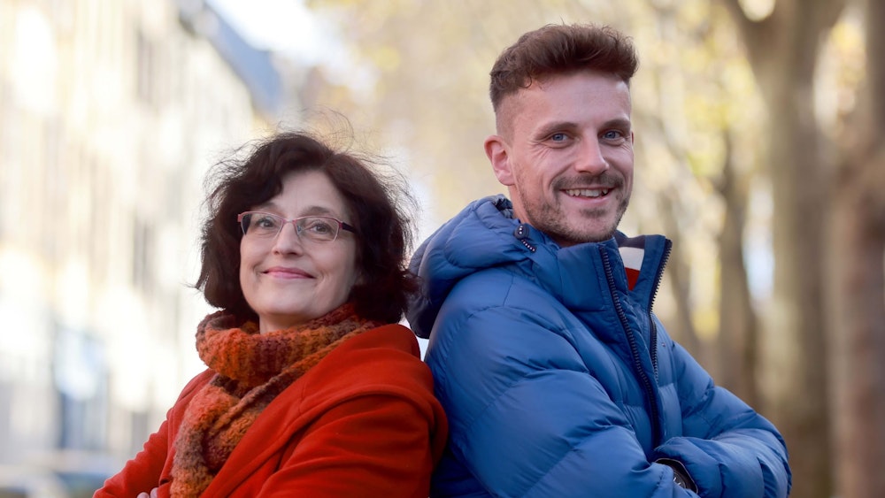Claudia Walther mit roter Jacke und Florian Schuster in blauer Jacke. Beide lächeln Rücken an Rücken in die Kamera.