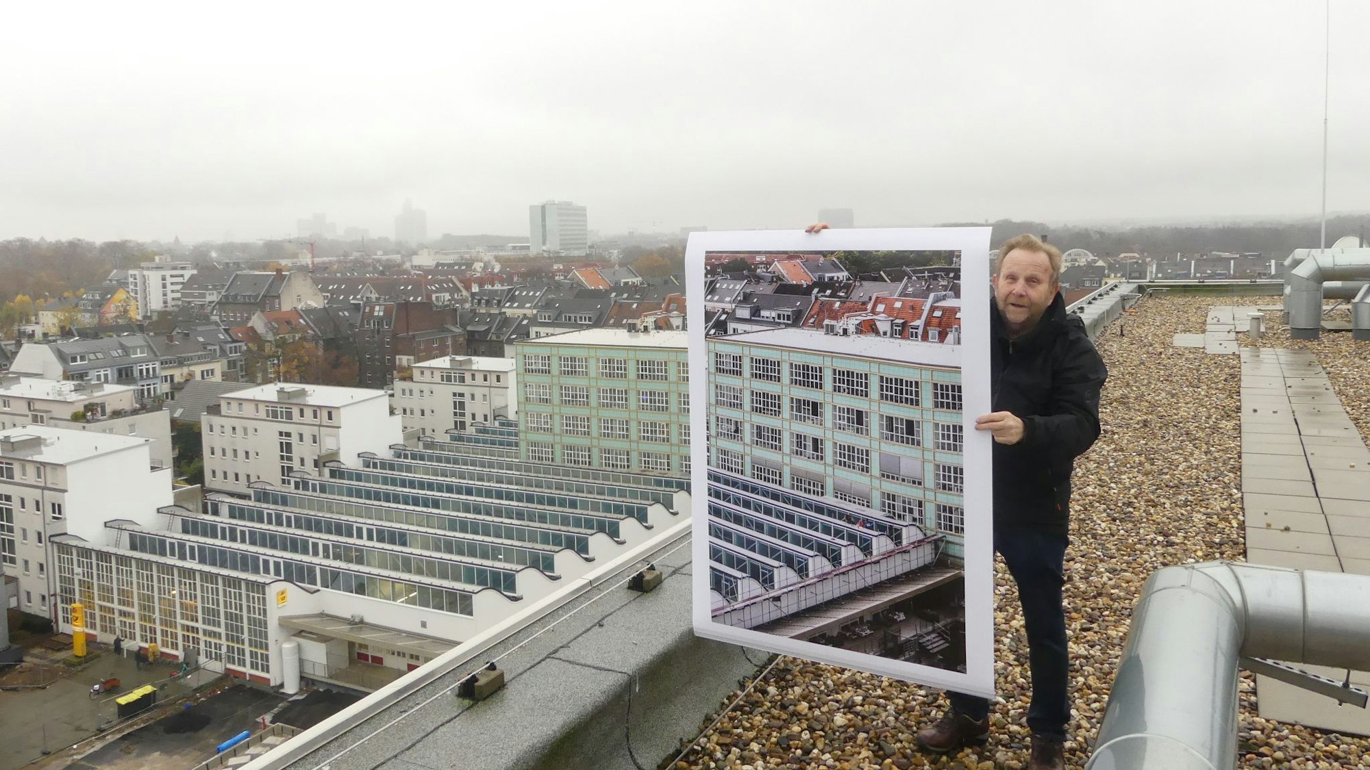 Fotograf Achim Bednorz auf dem 4711-Genäude in Ehrenfeld, während er eine seiner Fotografien in die Luft hält, die Stadt im Hintergrund.