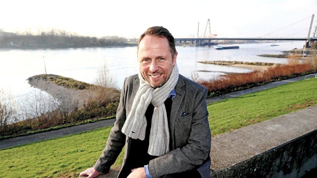 Uwe Richrath sitzt auf einer Mauer vor einer Böschung, die zum Rhein führt. Der Rhein ist groß im Hintergrund, ein Schiff ist zu sehen und die Leverkusener Rheinbrücke.