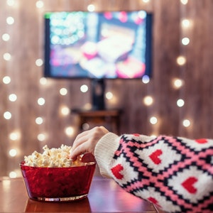 Eine Person greift in eine Schüssel voller Popcorn. Im Hintergrund sind ein Weihnachtsbaum, festliche Beleuchtung sowie ein laufender Fernseher zu sehen.