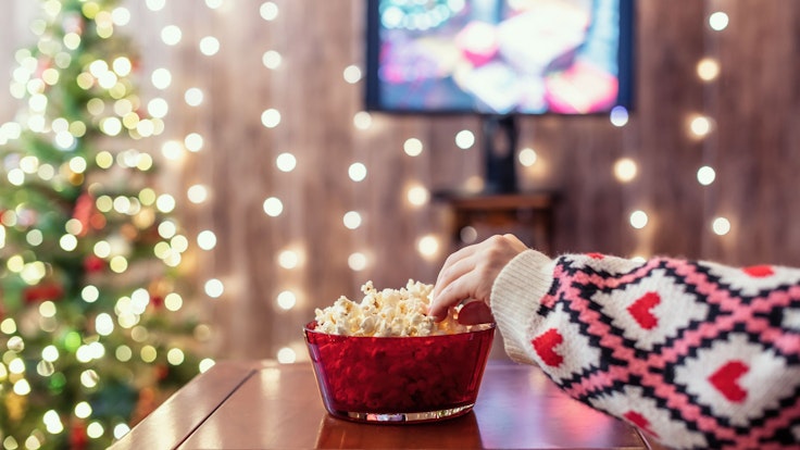 Eine Person greift in eine Schüssel voller Popcorn. Im Hintergrund sind ein Weihnachtsbaum, festliche Beleuchtung sowie ein laufender Fernseher zu sehen.