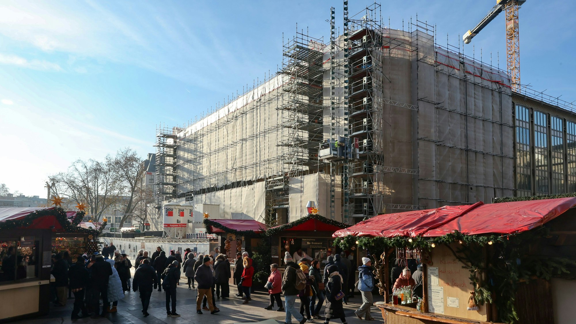 Das Bild zeigt das eingerüstete Dom-Hotel, davor stehen Buden des Weihnachtsmarktes auf dem Roncalliplatz.