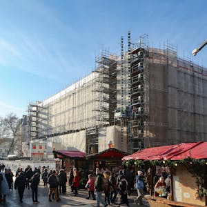 Das Bild zeigt das eingerüstete Dom-Hotel, davor stehen Buden des Weihnachtsmarktes auf dem Roncalliplatz.