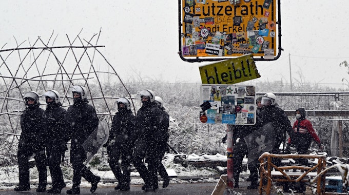 Polizisten stehen bei dem mit Stickern zugeklebten Ortsschild zum Eingang von Lützerath. Um sie herum liegt Schnee.
