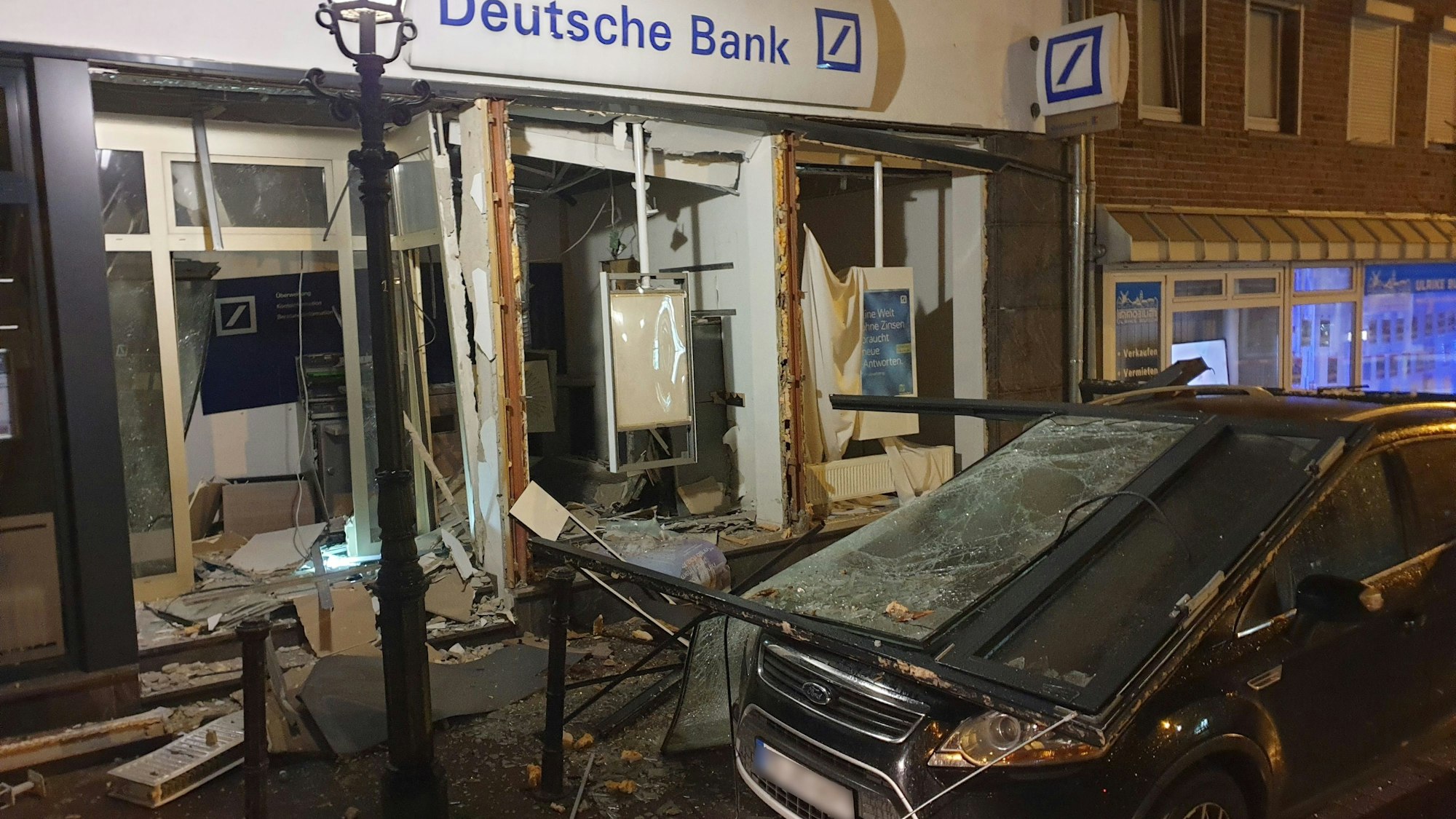 Ein stark beschädigtes Auto steht vor einer Bankfiliale, die durch eine Geldautomatensprengung verwüstet ist. Unbekannte hatten den Geldautomaten gesprengt. Anschließend flüchteten sie in einem dunklen Auto.