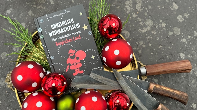 Das Buch „Unheimlich weihnachtlich – Böse Geschichten aus dem Bergischen Land“ liegt in einer Schale mit Weihnachtskugeln - und drei Messern.