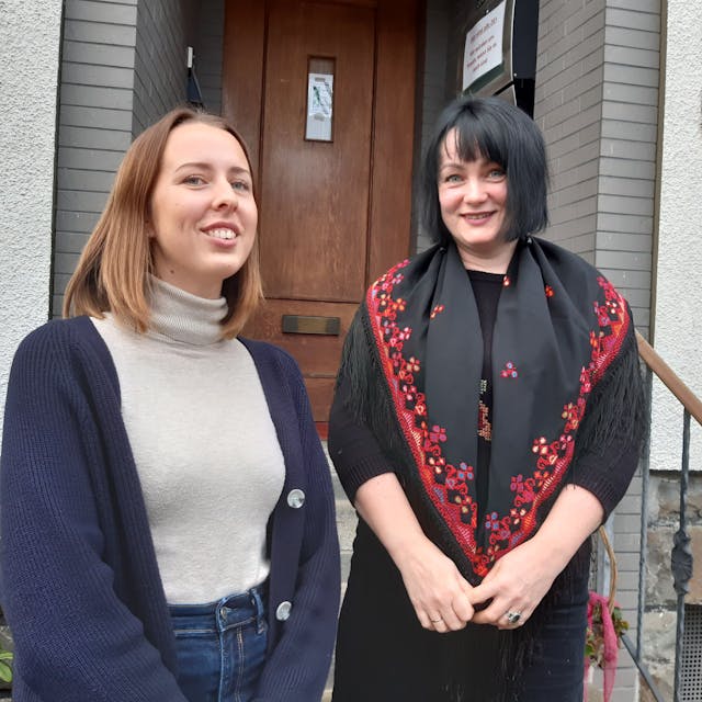 Zwei Frauen stehen vor einer Tür mit der Hausnummer fünf und lächeln in die Kamera.