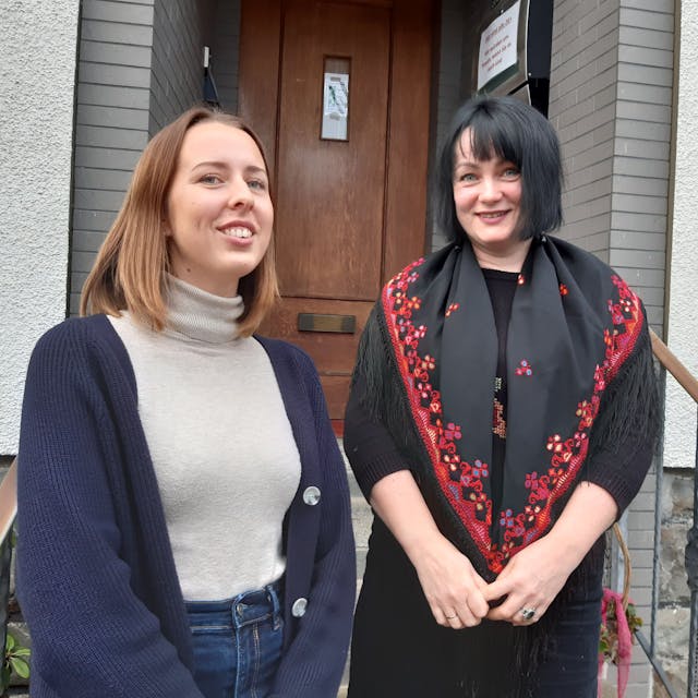 Zwei Frauen stehen vor einer Tür mit der Hausnummer fünf und lächeln in die Kamera.