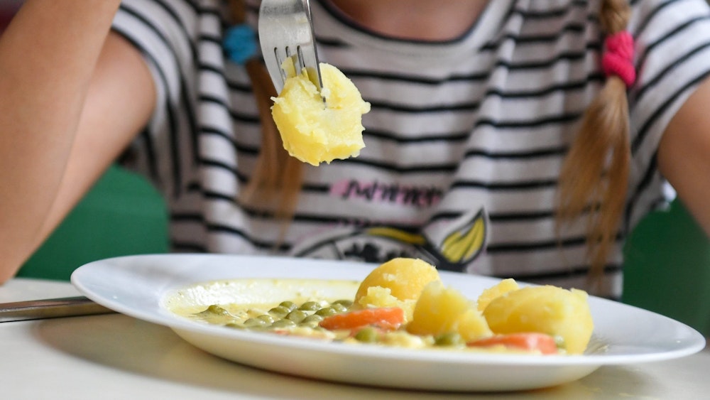 Eine Schülerin sitzt in einer Schulmensa vor einem Teller mit Kartoffeln, Erbsen und Möhren.