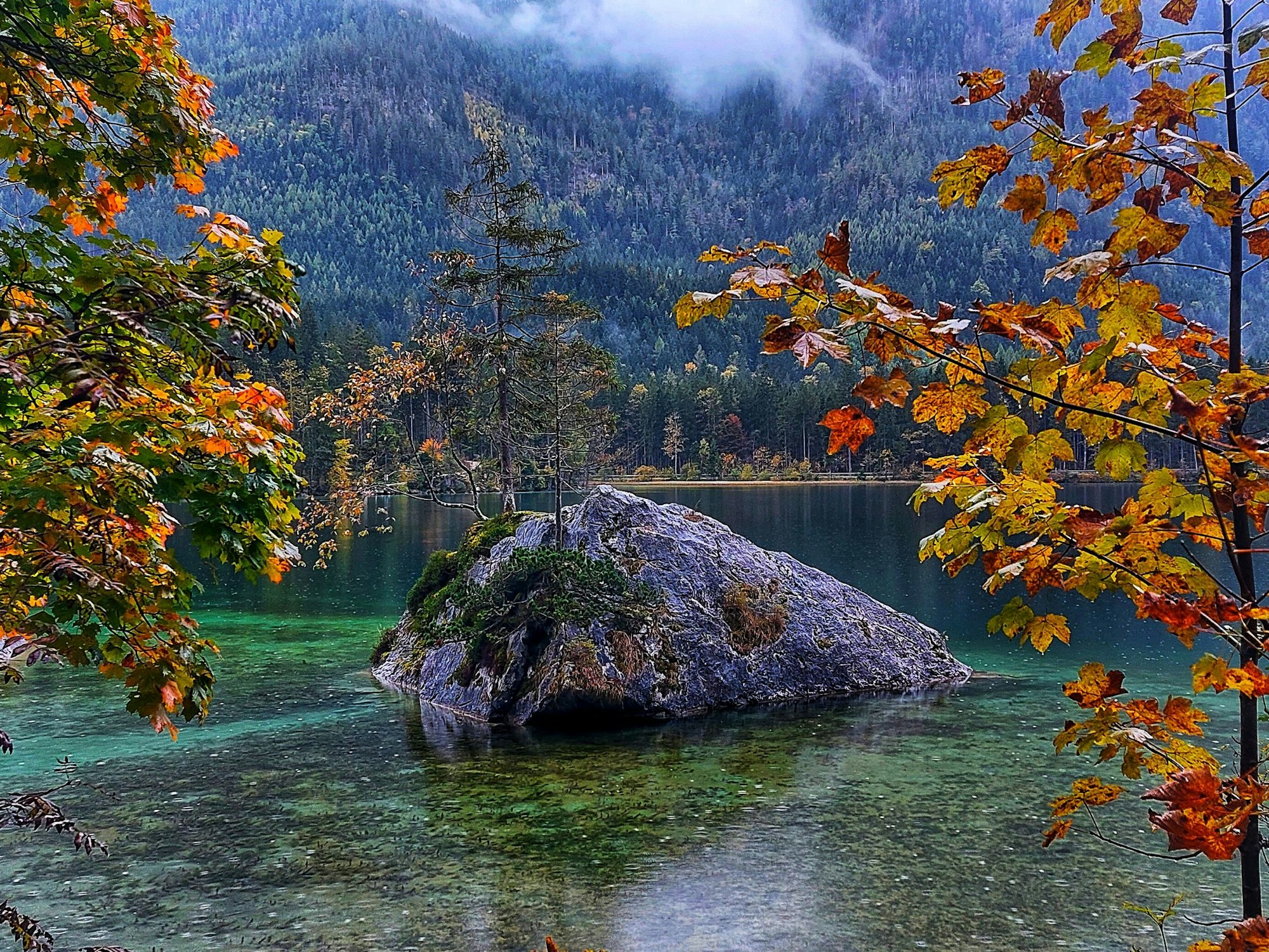 Ein farbenprächtiger See, in der Mitte liegt ein Fels mit Baum darauf, hinten ein Wald, rechts und links im Vordergrund bunte Herbstbäume.