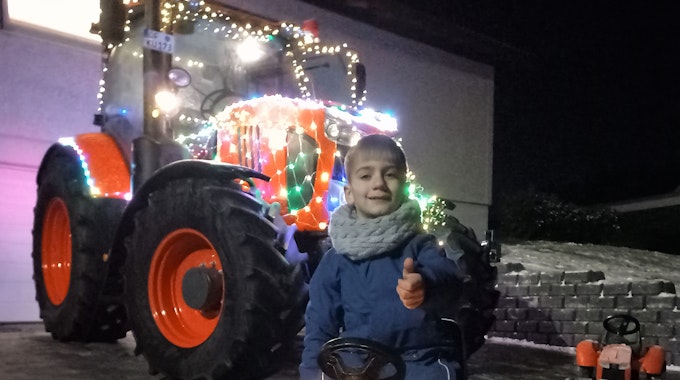 Blick auf einen Jungen auf einem mit Lichtern geschmückten Spieltraktor. Im Hintergrund ein mit Lichtern geschmückter großer Traktor.