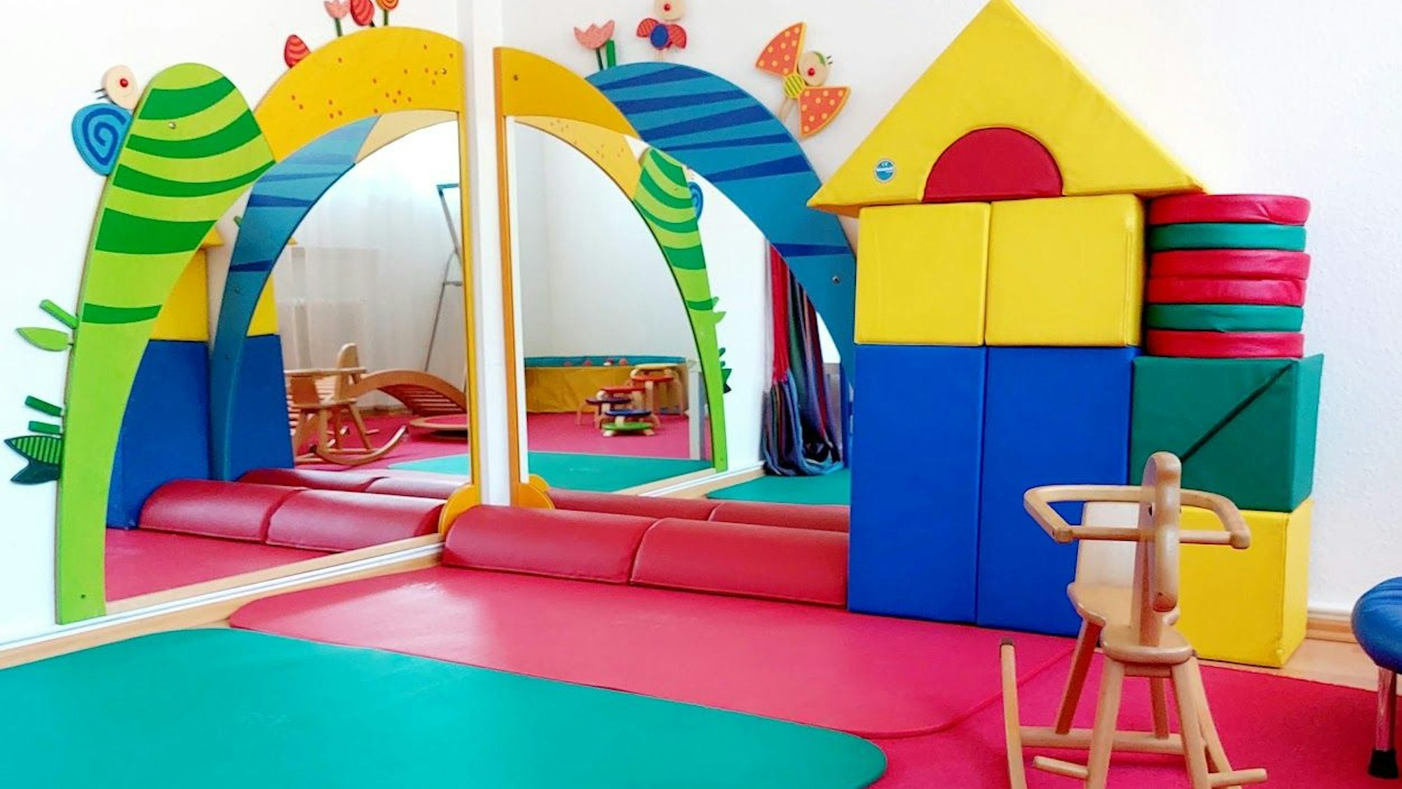 Ein Raum für Kinder mit bunten Matten und überdimensonal großen Bauklötzen, einem Schaukelpferd und großen Spiegeln.