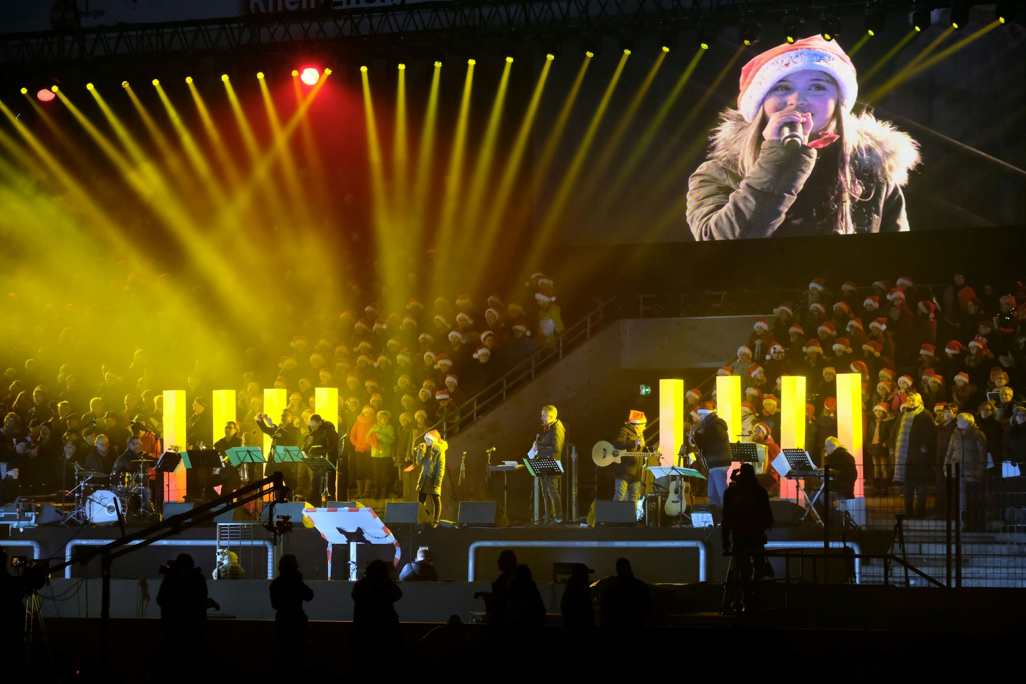 Musiker stehen vor einem Chor mit Weihnachtsmützen auf der Südtribüne des Kölner Stadions im Scheinwerferlicht. Auf einer großen Videoscreen ist ein Mädchen mit Nikolausmütze zu sehen, das singt.