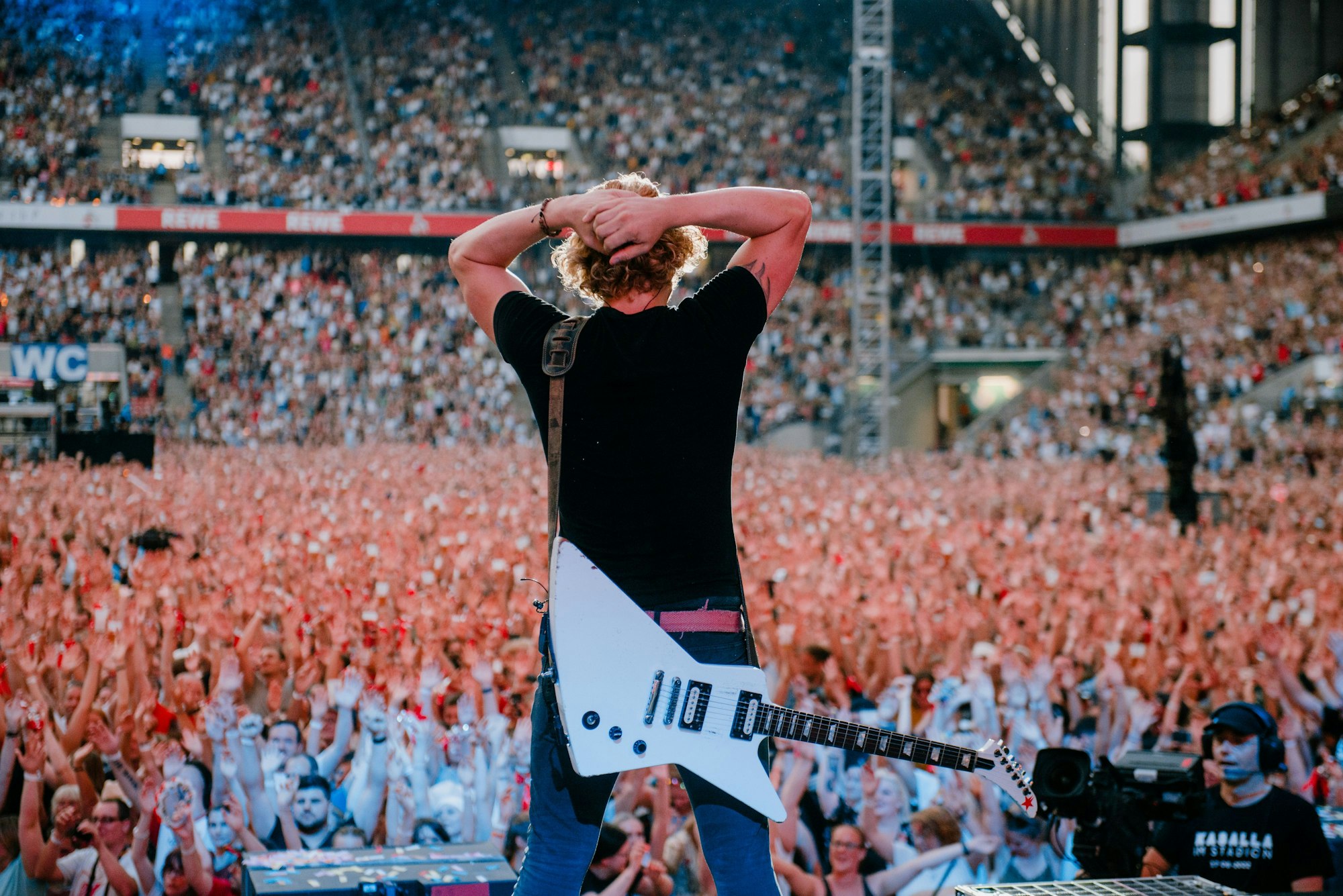 Beim Kasalla-Konzert im Stadion steht  Gitarrist Flo Peil mit den Händen überm Kopf vor der jubelnden Menge