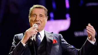 Udo Jürgens bei einem Live-Auftritt im Anzug. Er singt in ein Mikrofon und hebt die linke Hand.
