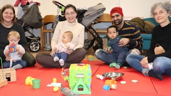 Drei Eltern sitzen mit ihren Babys und einer Diplom-Heilpädagogin auf knallroten Matten, vor ihnen liegt allerhand Spielzeug.