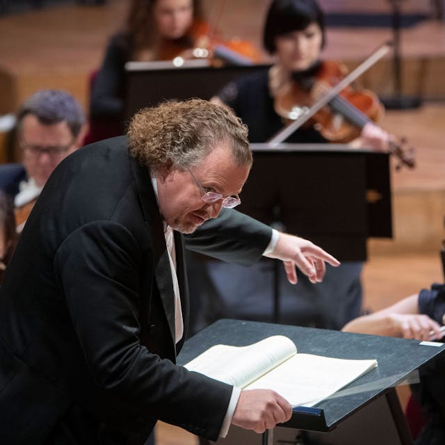 Der französische Dirigent Stéphane Denève beugt sich über sein Pult, im Hintergrund sind Musiker zu sehen.