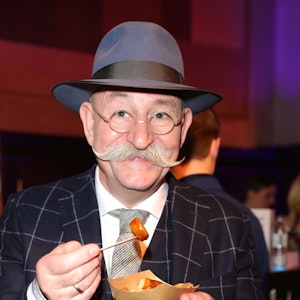 Horst Lichter lächelt beim Deutschen Fernsehpreis in einem Anzug in die Kamera und isst Pommes mit Currywurst.