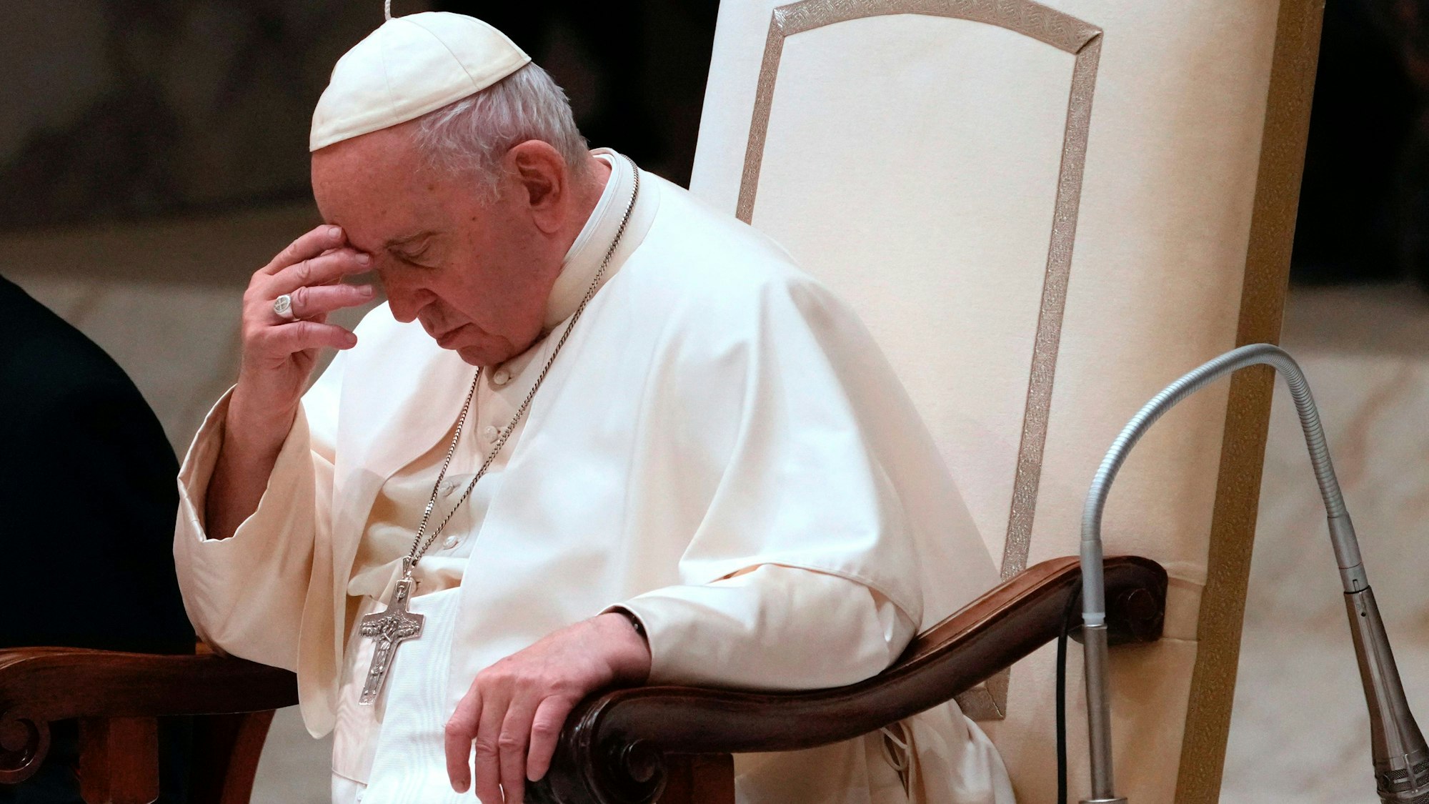 Papst Franziskus sitzt in einem Stuhl und fasst sich an den Kopf. Um seinen Hals hängt ein silbernes Kreuz.