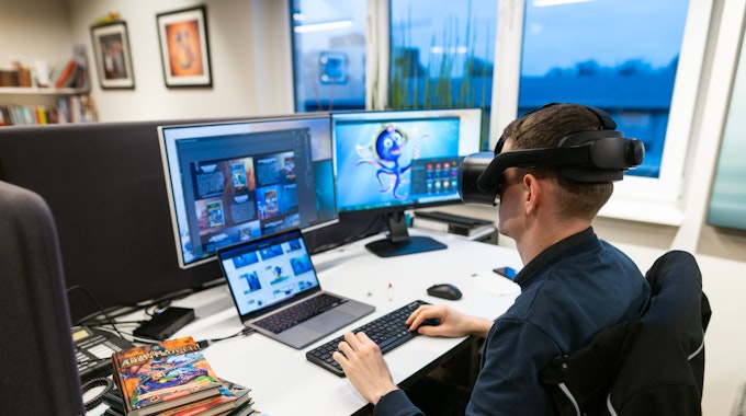Ein Mann sitzt mit VR-Brille an einem Arbeitsplatz mit mehreren PC-Bildschirmen.