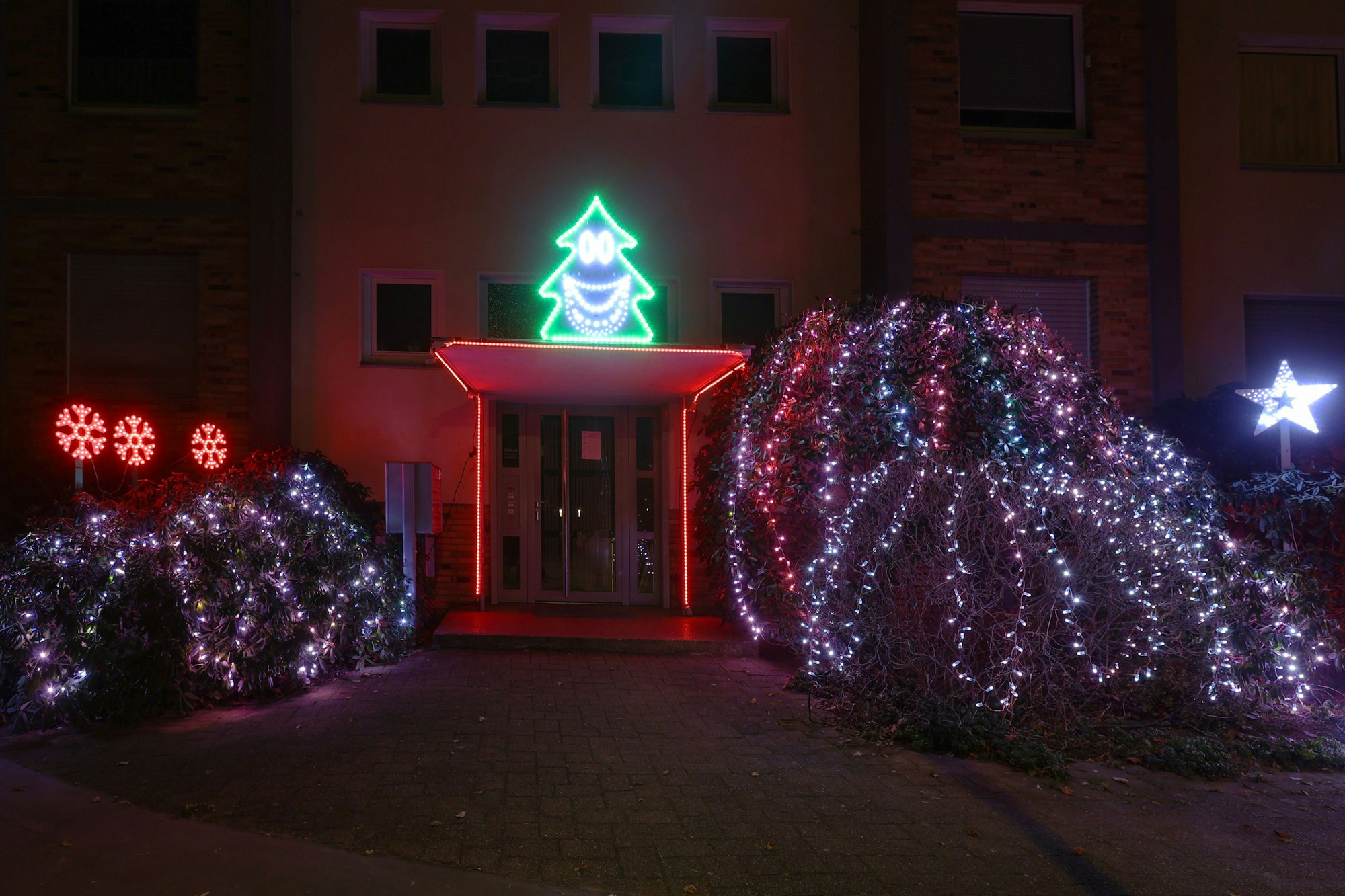 Vor einem Hauseingang hängen Lichterketten in den Sträuchern. Links sind drei rot leuchtende Eiskristalle zu sehen. Über der Tür leuchtet die Silhouette eines Weihnachtsbaums mit einem Gesicht.