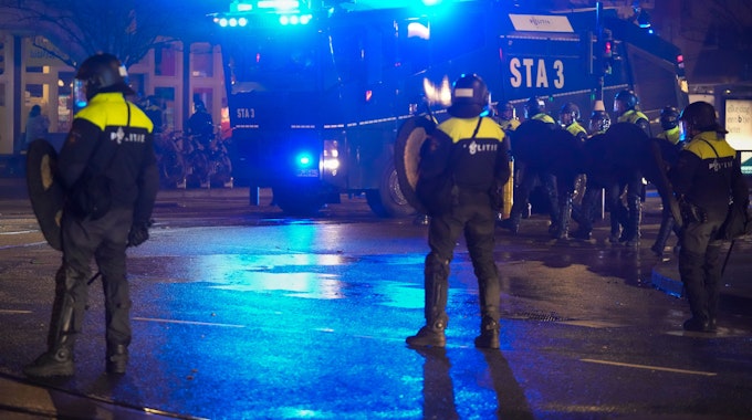 Die niederländische Polizei steht bei Dunkelheit auf einer großen Straße in Amsterdam, im Hintergrund steht ein Wasserwerfer der Bundespolizei aus Sankt-Augustin.