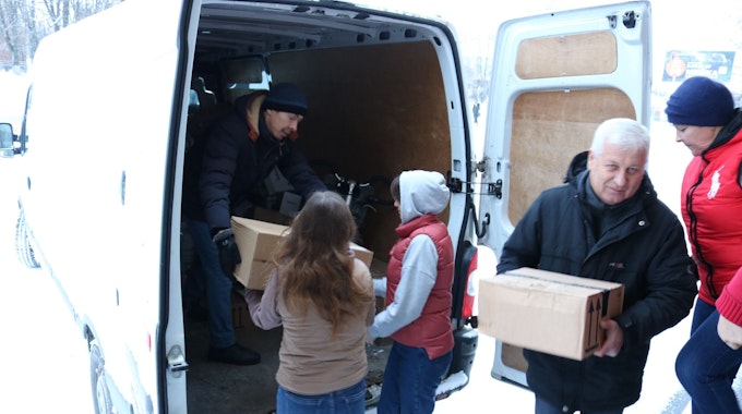 Männer, Frauen und Kinder helfen beim Entladen eines weißen Transporters, sie reichen sich Kartons an.