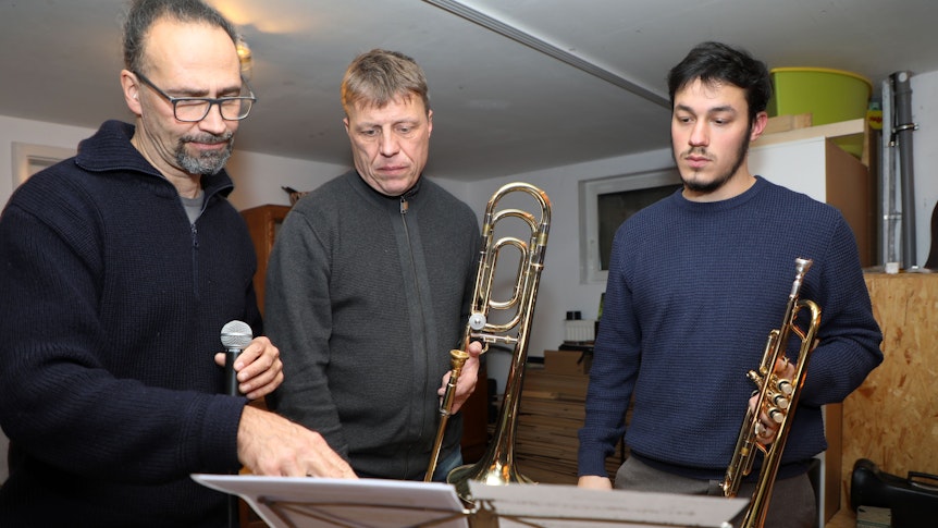 Eddi Hüneke (Mikro in der Hand), Clemens Tewinkel (Posaune) und Jonathan Anand (Trompete) stehen vor einem Notenständer und diskutieren über die Noten.