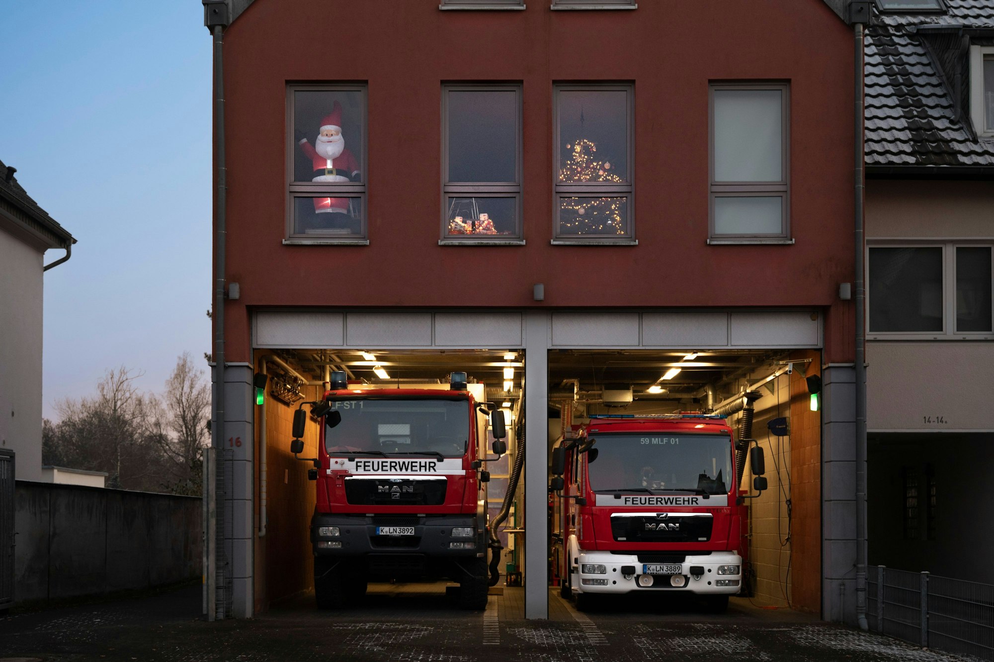 Das Gerätehaus ist mehrstöckig. In den Fenstern im ersten Stock sind ein leuchtender Weihnachtsmann sowie ein Tannenbaum zu sehen. In den zwei Fahrzeughallen im Erdgeschoss stehen zwei Feuerwehrfahrzeuge.