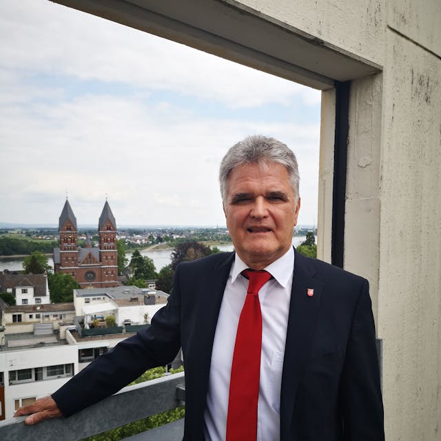 Erwin Esser in schwarzem Anzug und roter Krawatte. Im Hintergrund ist die Stadt Wesseling zu sehen.