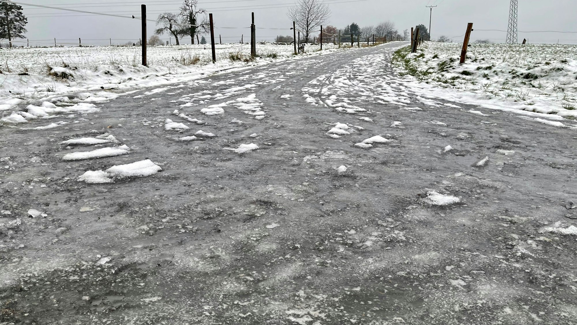 Eine dicke Eisschicht liegt auf der Straße. Felder sind mit Zäunen abgetrennt.