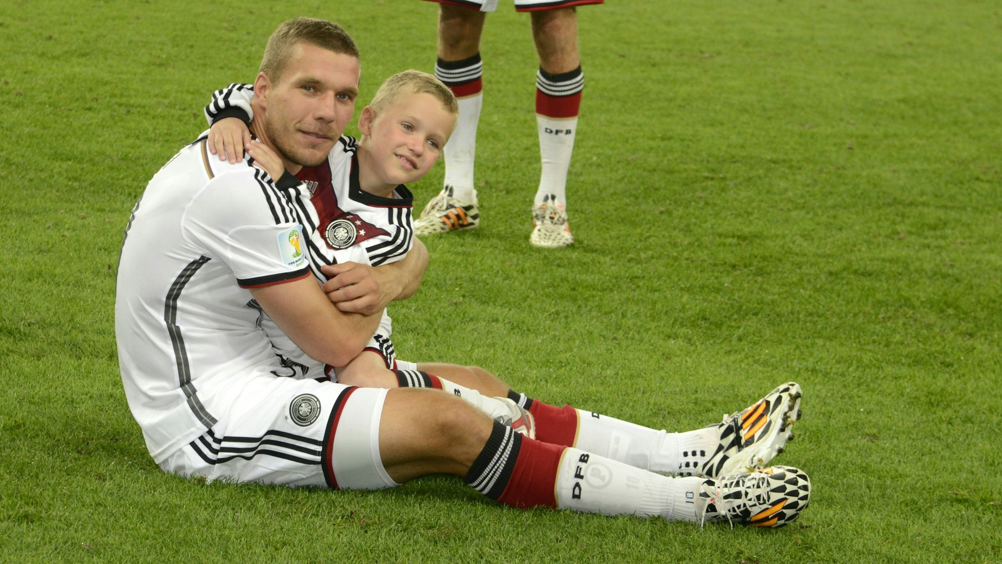Lukas Podolski sitzt mit seinem Sohn auf dem Arm auf dem Rasen.