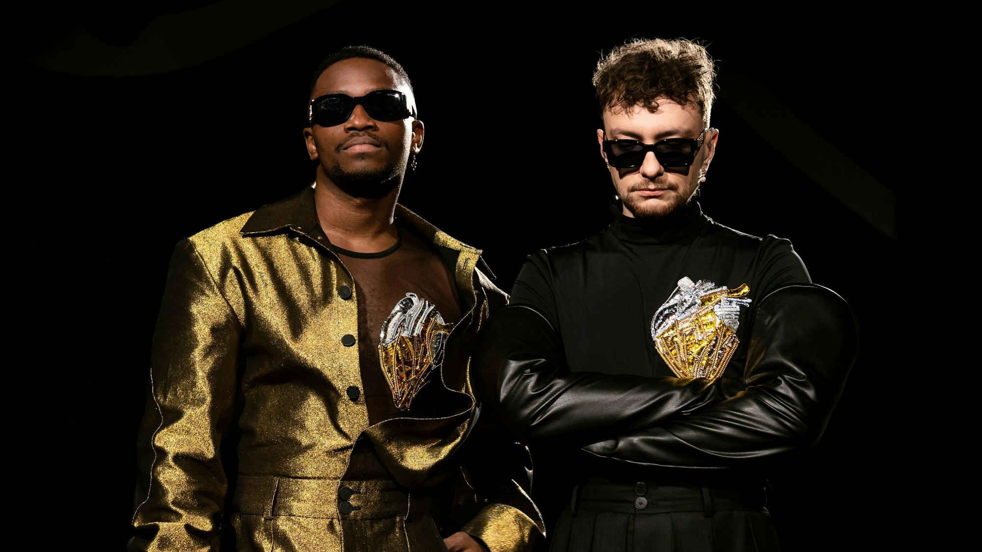 Jeffery Kenny (l) und Andrij Guzuljak von der Band TVORCHI in einer U-Bahn-Station. Sie tragen goldene bzw. schwarze Bühnenoutfits und dunkle Sonnenbrillen.