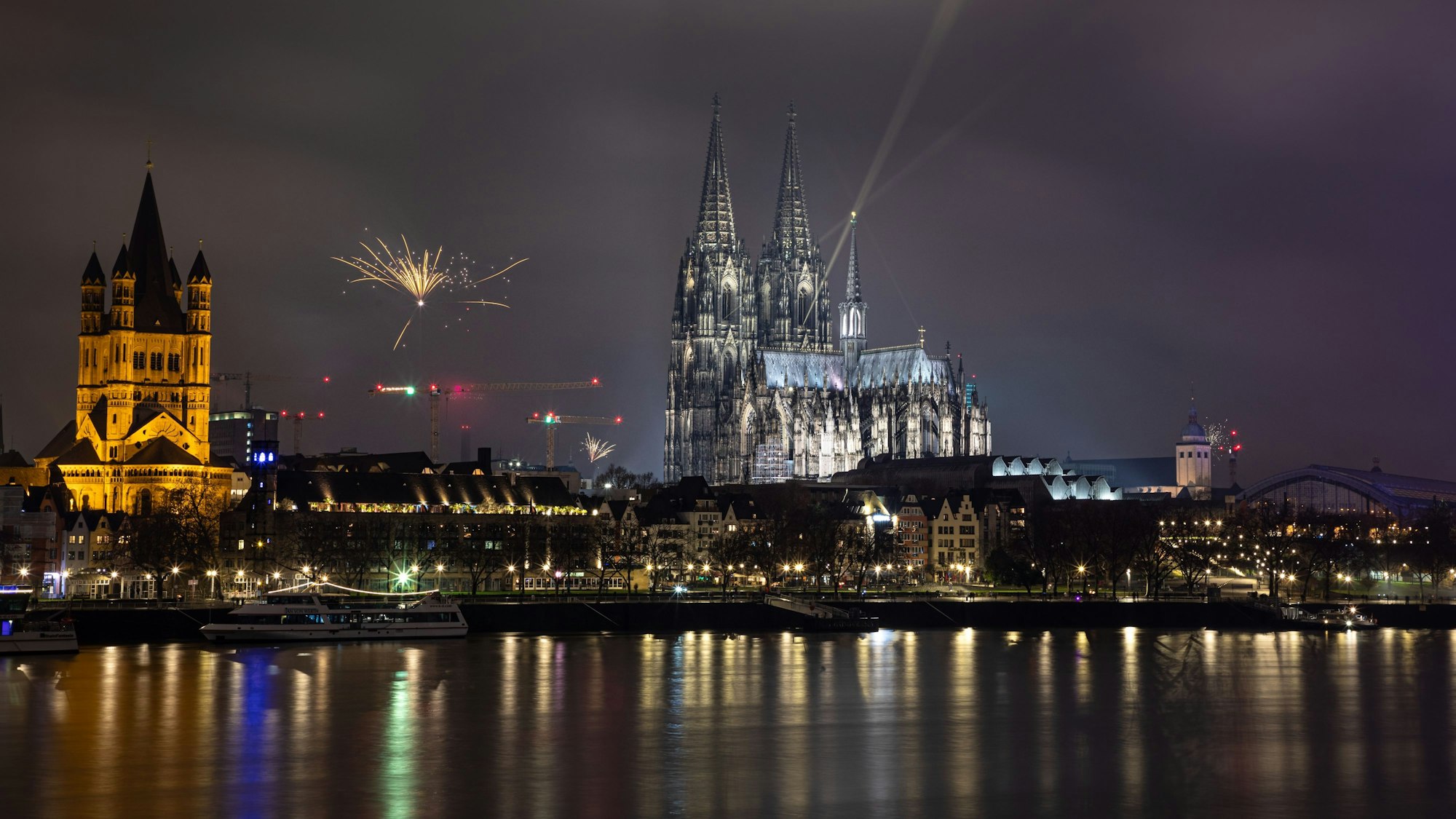 Die Nachtaufnahme zeigt das Kölner Panorama mit dem Rhein im Vordergrund und den Kirchen Kölner Dom sowie Groß St. Martin. Am Himmel explodiert eine einzelne Silvesterrakete.
