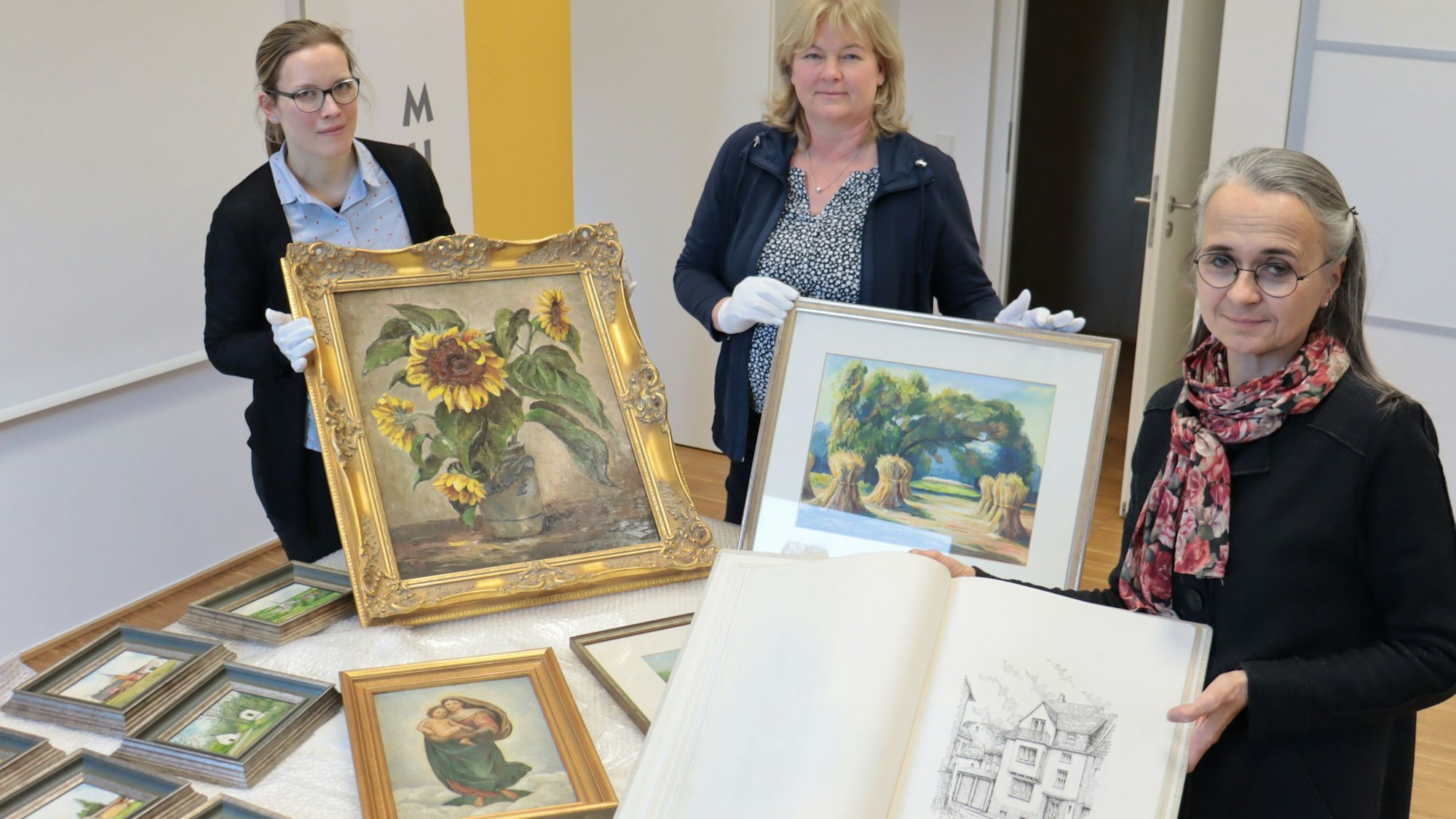 Zeichnungen und Gemälde des Euskirchener Malers Willy Loos, die Enkelin Iris Pütz an Leiterin Heike Lützenkirchen und deren Mitarbeiterin Jennifer Kirchhoff überreicht, sind auf einem Tisch ausgebreitet.
