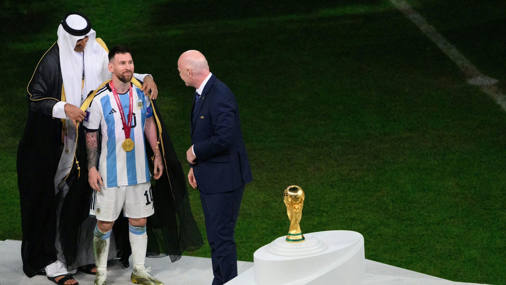 Der Emir von Katar, Scheich Tamim bin Hamad Al Thani (l), legt Argentiniens Lionel Messi vor Übergabe des WM-Pokals vor Fifa-Präsident Gianni Infantino ein Bischt, ein traditionelles arabische Kleidungsstück an.