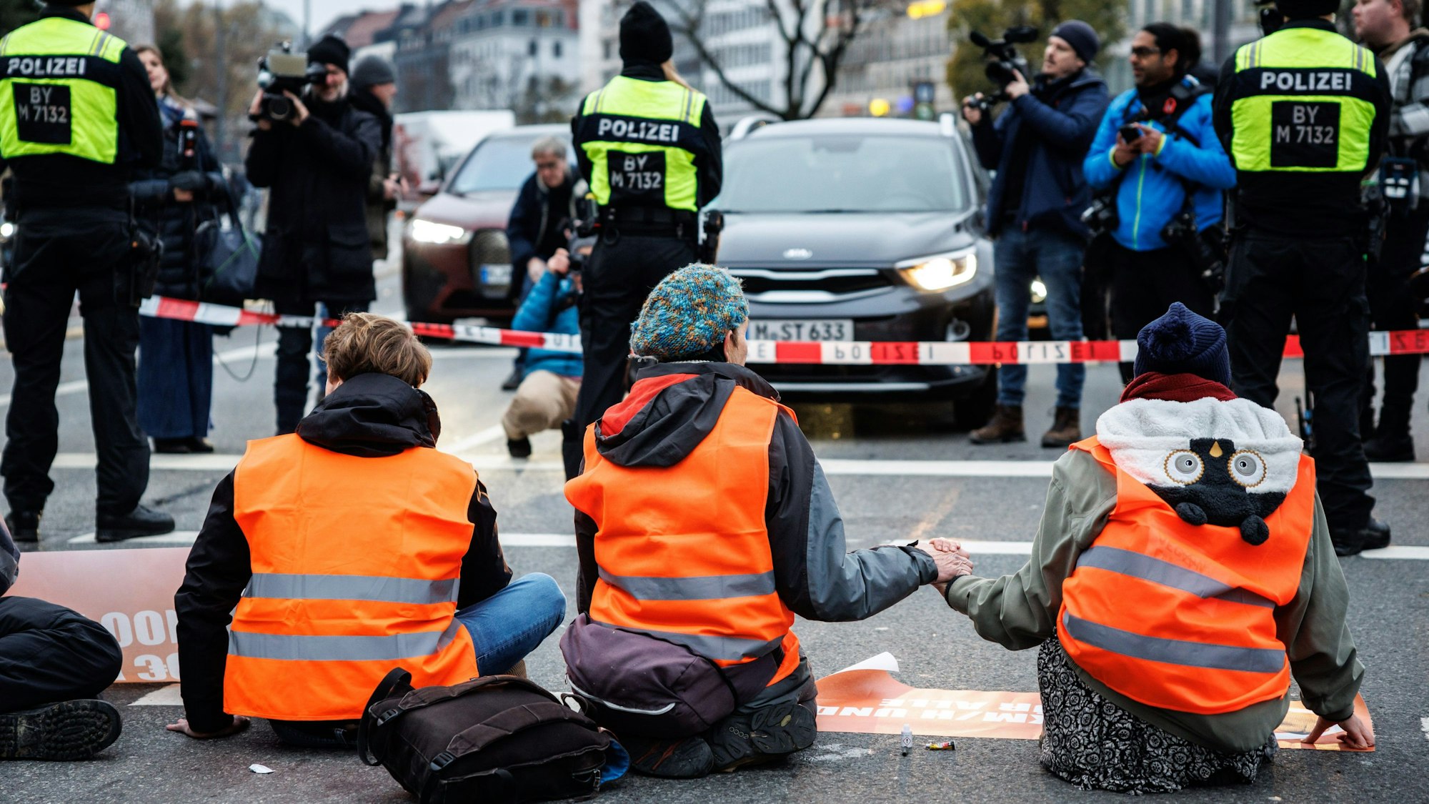 Drei Jugendliche in orangefarbigen Warnwesten blockieren sitzend eine Fahrbahn. Drei Polizisten sowie Absperrband schützen die Klimaaktivisten vor Autos und Fotografen.