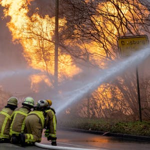 Feuerwehrleute arbeiten daran, das brennende Gasleck in Duisburg zu löschen. Ein Feuer und Rauchschwaden sind zu sehen, im Vordergrund die Feuerwehrleute.