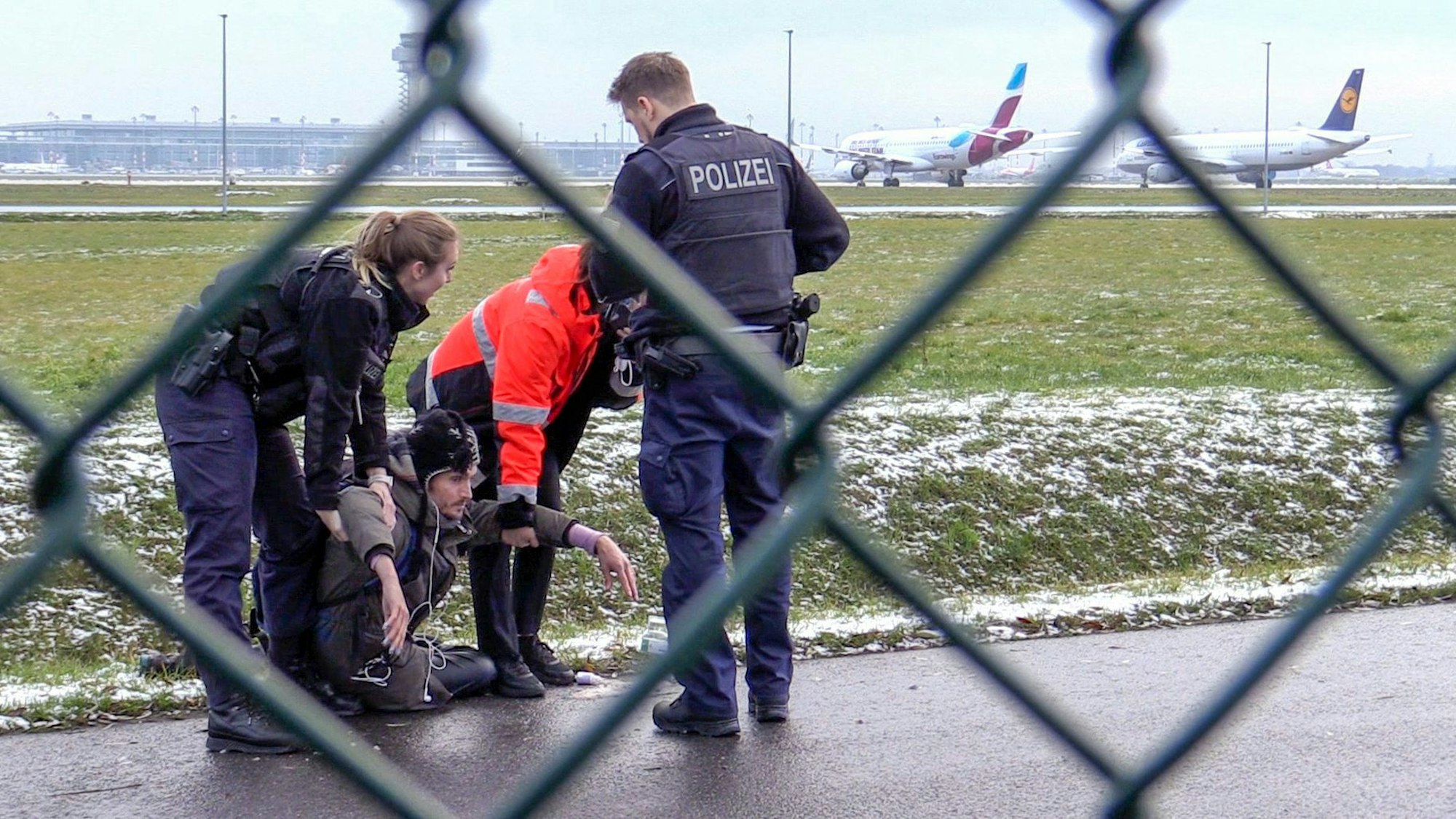 Eine Polizistin und ein Mitarbeiter des Berliner Flughafens versuchen, einen am Boden sitzenden Klimaaktivisten anzuheben. Ein zweiter Polizist steht vor der Gruppe. Im Hintergrund sind zwei Flugzeuge und das Flughafengebäude zu sehen, im Bildvordergrund ein Maschendrahtzaun.