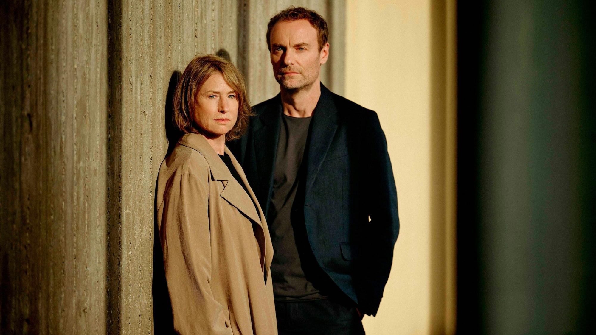 Die Schauspieler Corinna Harfouch als Susanne Bonard und Mark Waschke als Kriminalhauptkommissar Robert Karow bei den Dreharbeiten für den ersten gemeinsamen Berliner „Tatort“. Sie lehnen an einer Wand und schauen ernst.