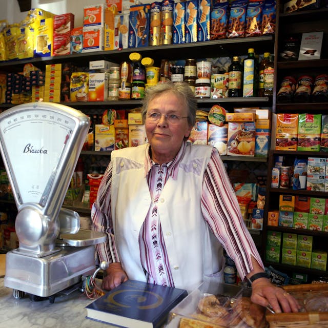 Eine ältere Dame mit Brille steht hinter einer Ladentheke, hinter ihr sind Regale mit Kaffee, Tee und anderen Lebensmitteln zu sehen, eine altmodische Waage steht neben.&nbsp;